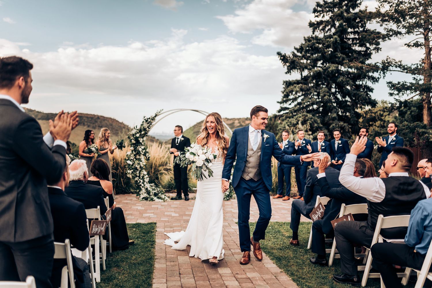 Wedding party walks down brick path aisle during outdoor ceremony with mountain backdrop.