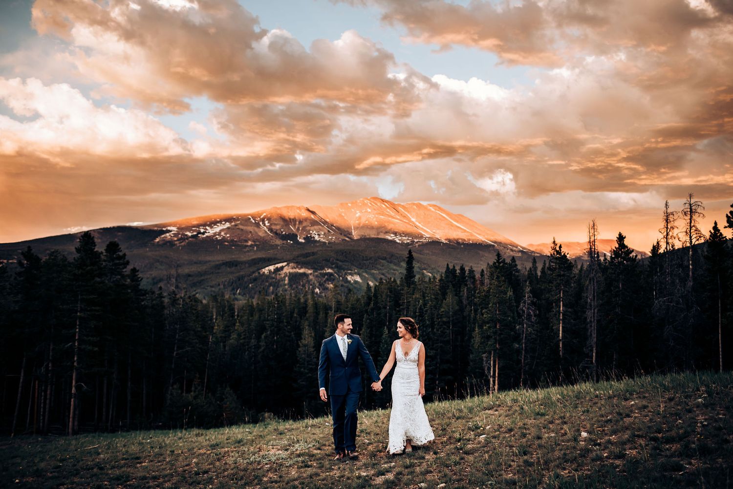 Newlyweds stand together on hillside with snow-capped mountains and orange sunset sky.