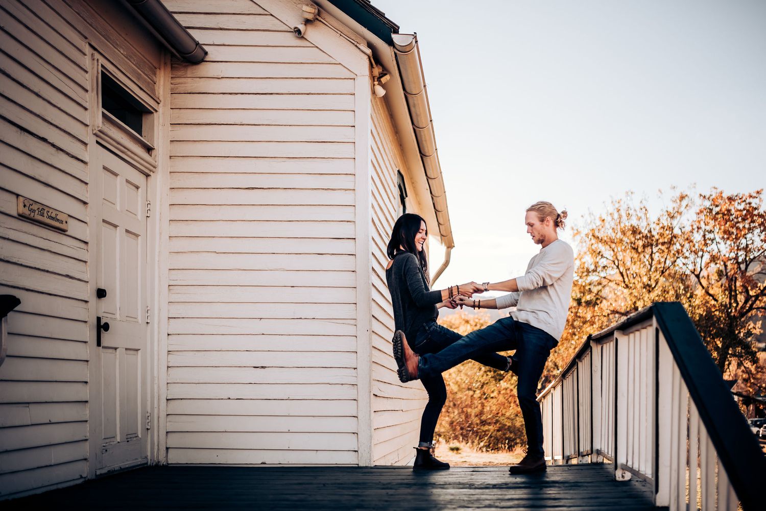 A playful couple balances on porch railings of a white house.