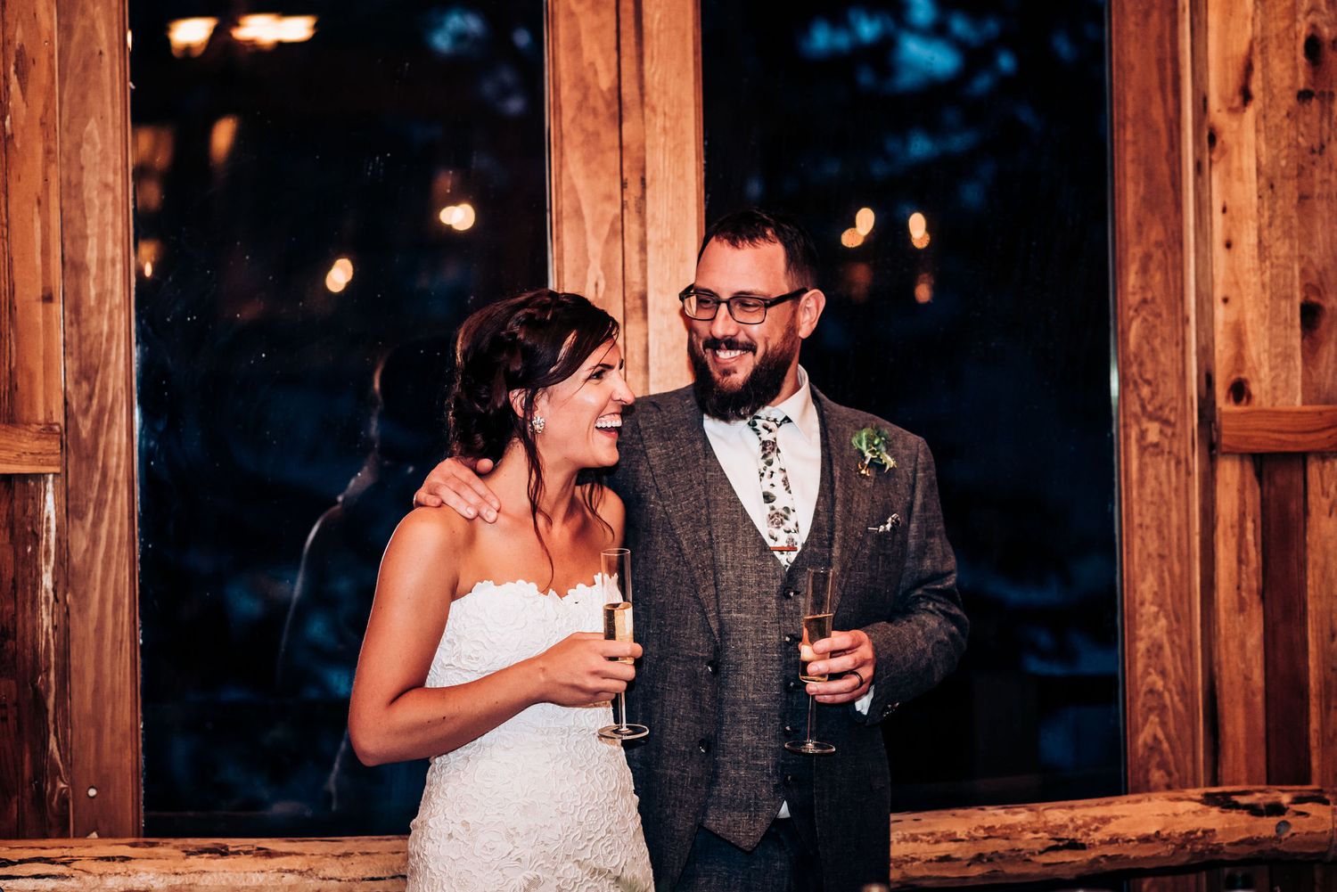 Couple shares drinks and laughter at rustic wooden lodge during evening reception.