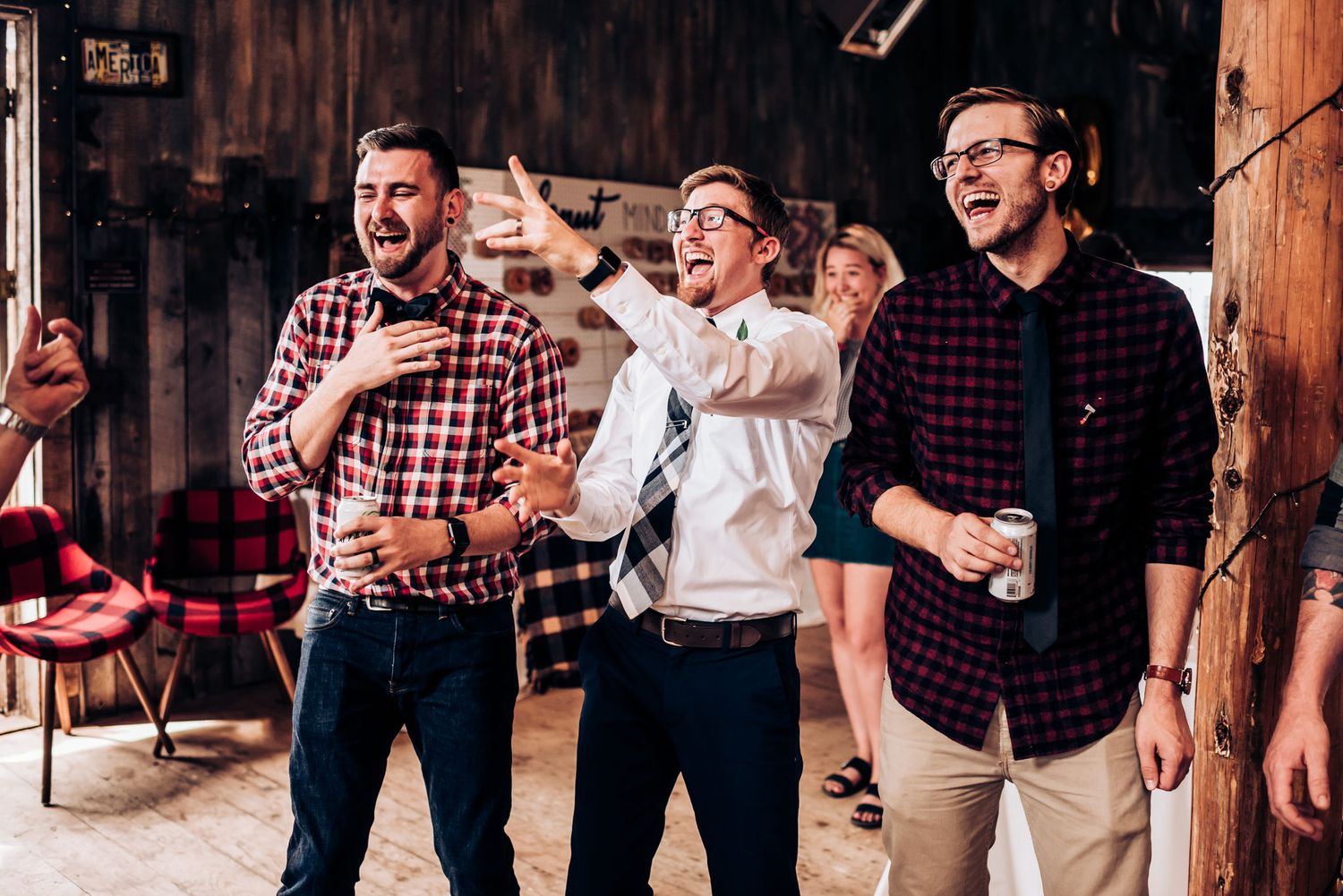 Three guests in plaid shirts celebrate at a rustic barn event with drinks in hand.