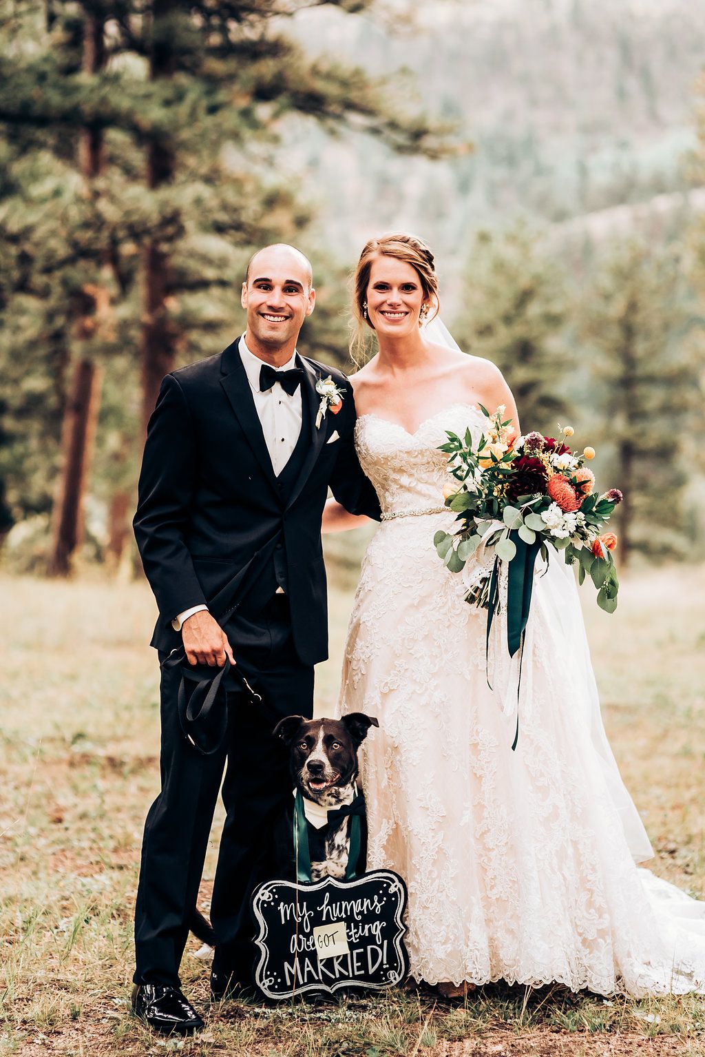 Black and white dog poses with newlyweds in rustic outdoor setting holding chalkboard wedding sign.