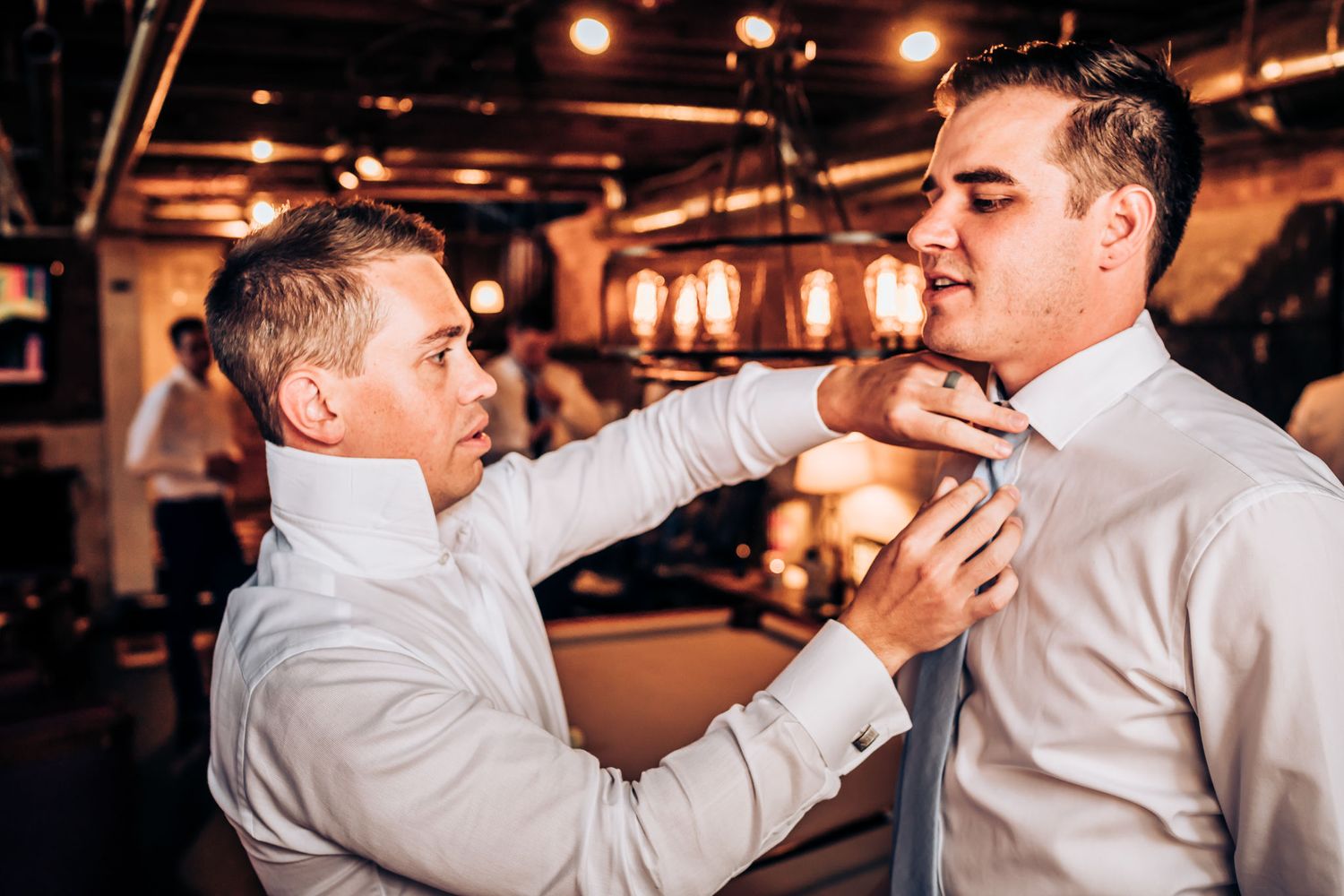 Two men in formal attire playfully adjusting each other's ties at indoor venue.