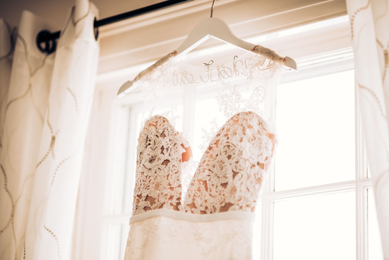 A detailed lace wedding dress bodice hangs in front of a bright window.