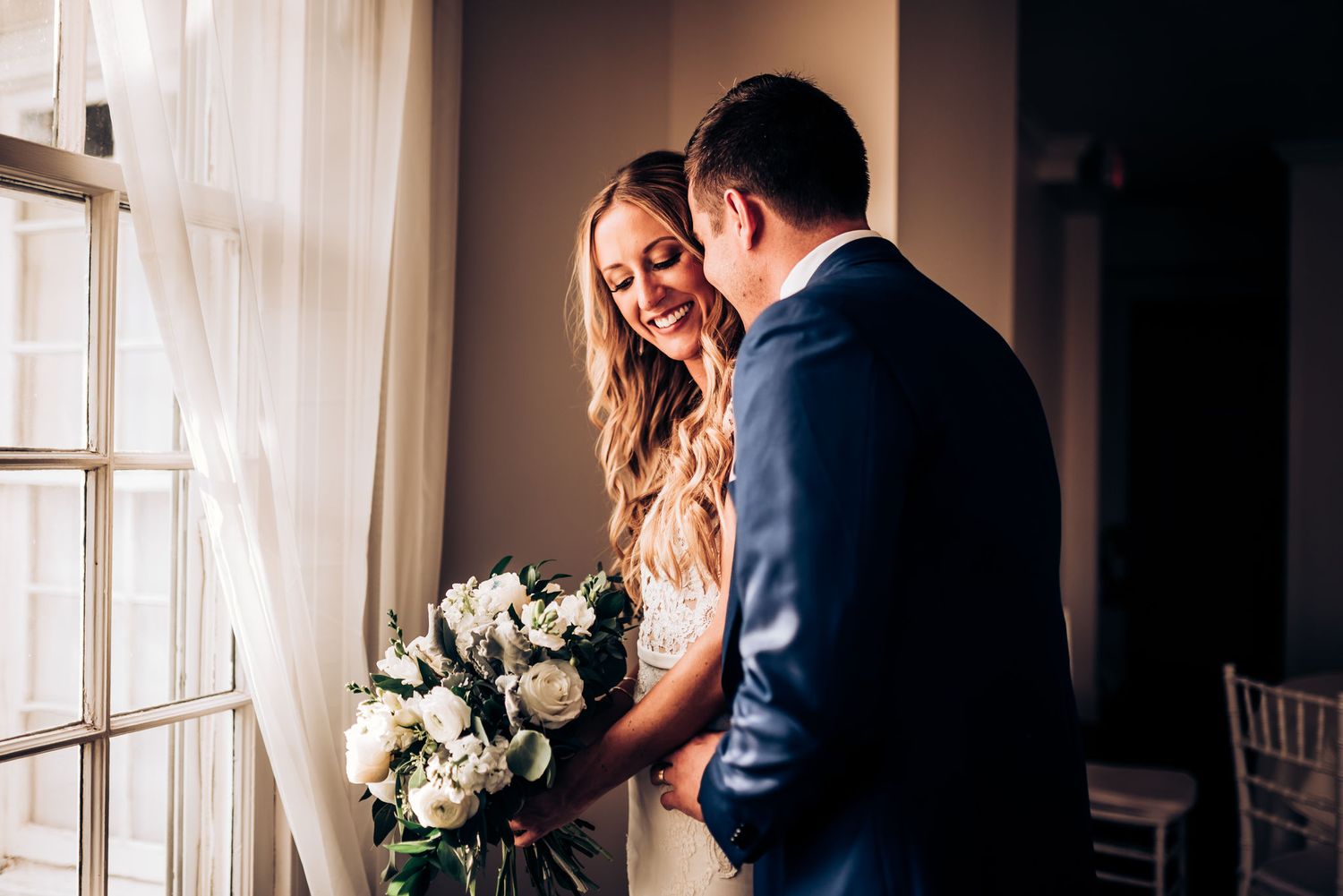 Intimate wedding moment captured by window with elegant white floral bouquet.