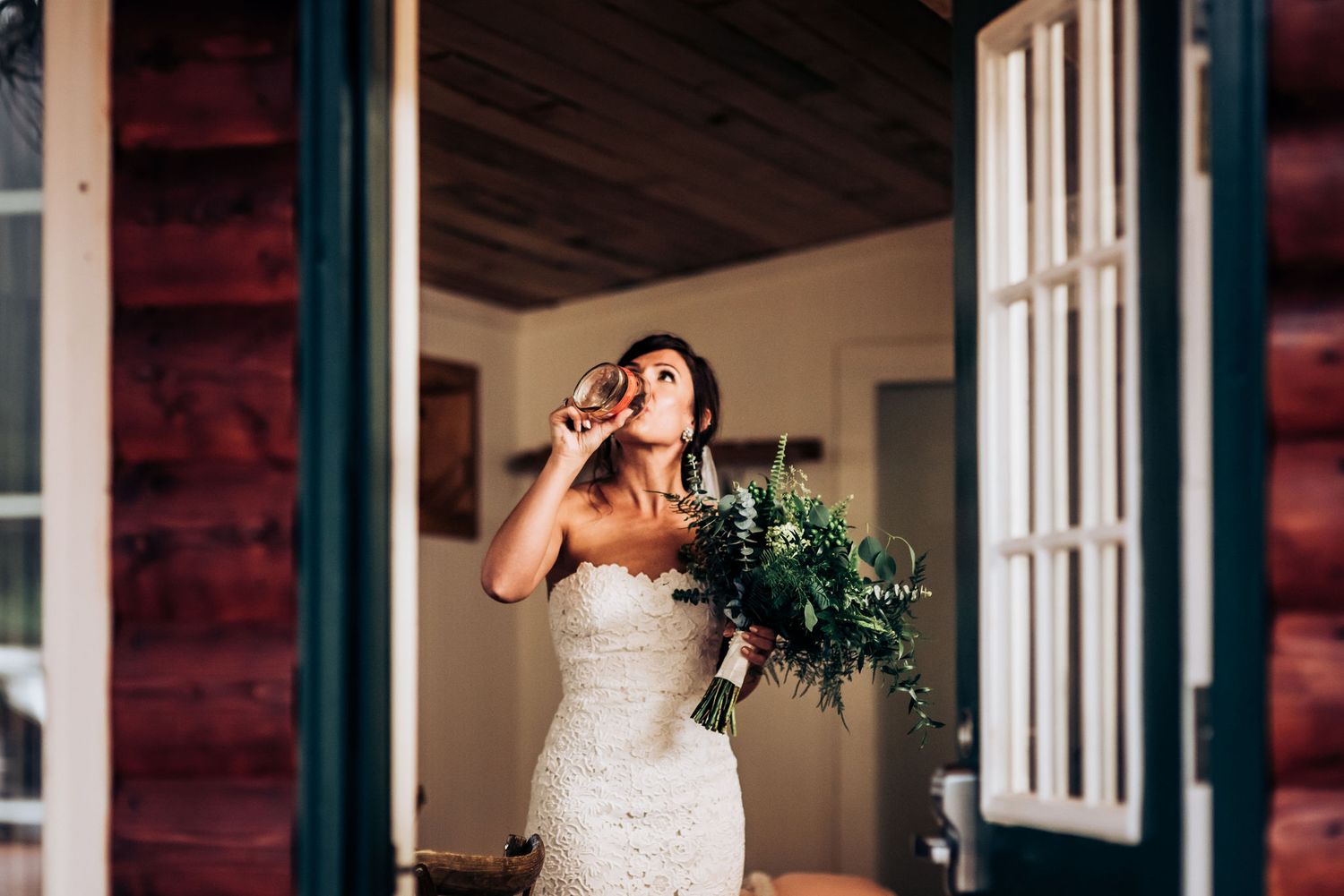 A bride in a lace dress holds a green bouquet while standing in front of an open door at a rustic venue.