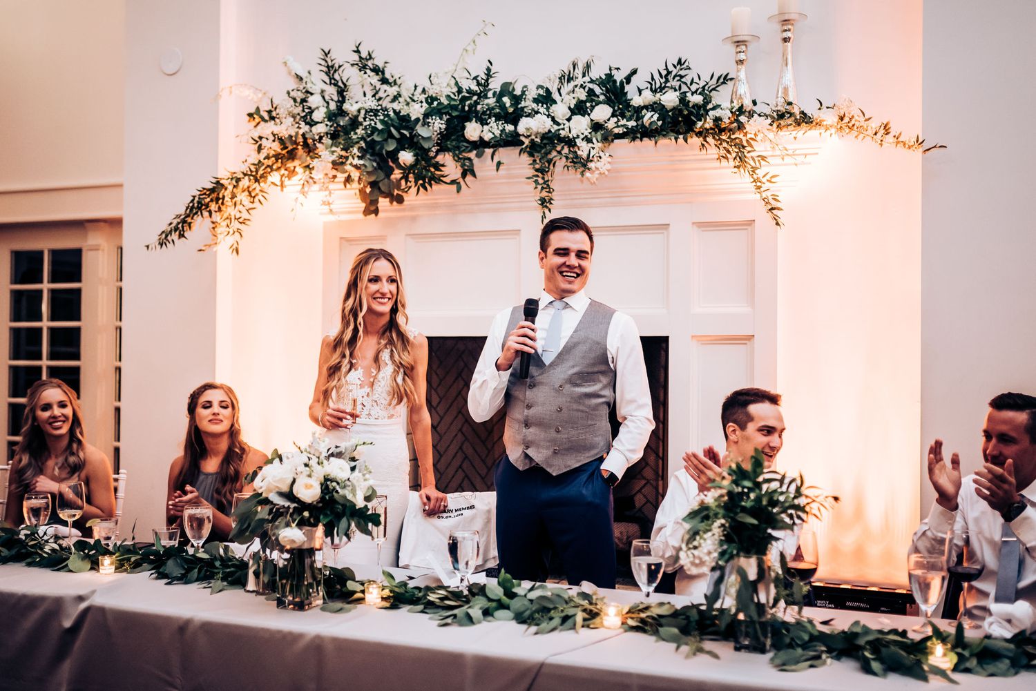 Couple stands at wedding reception head table decorated with elegant floral garlands and candlesticks.