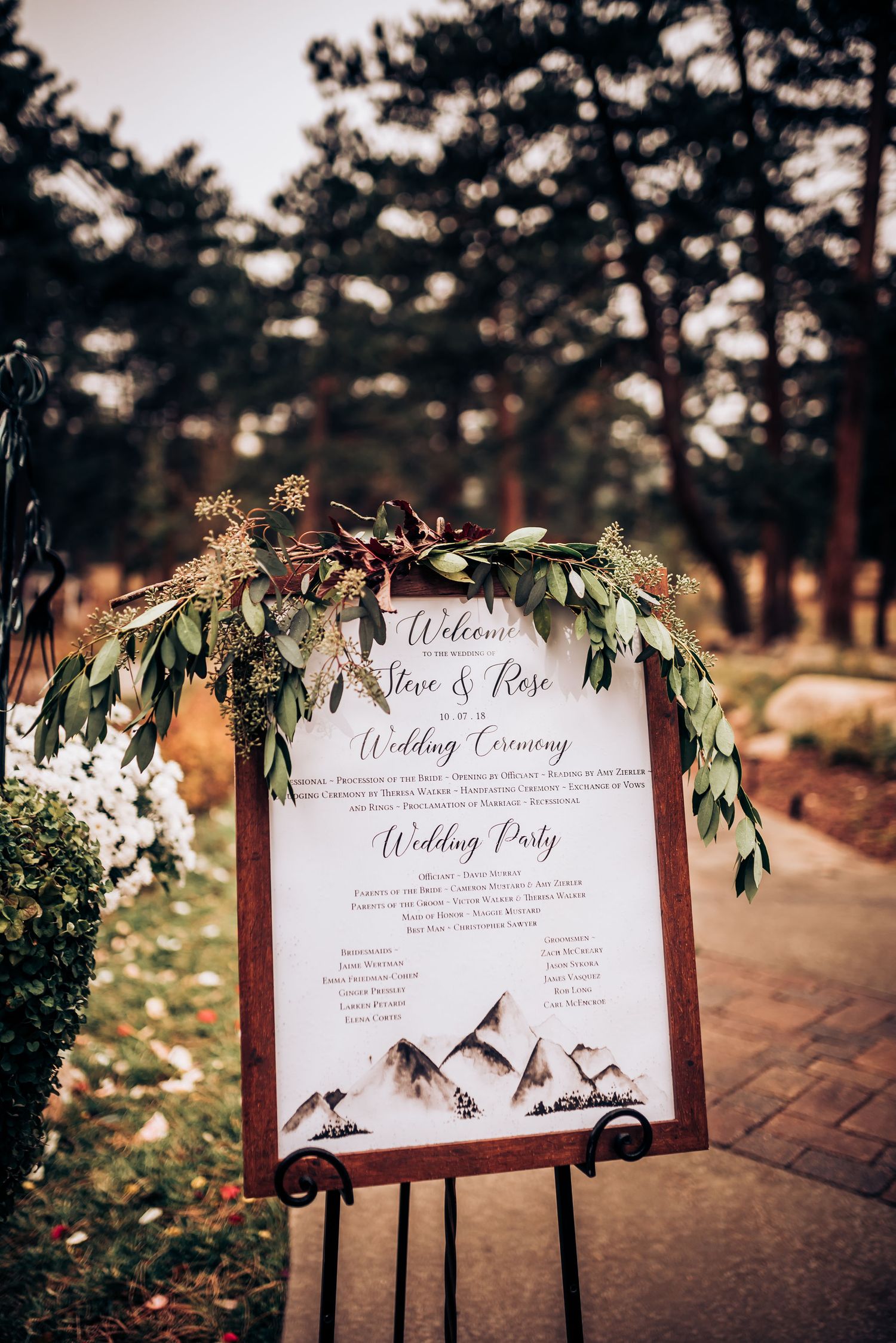Wedding ceremony program sign decorated with mountain graphics and greenery garland.