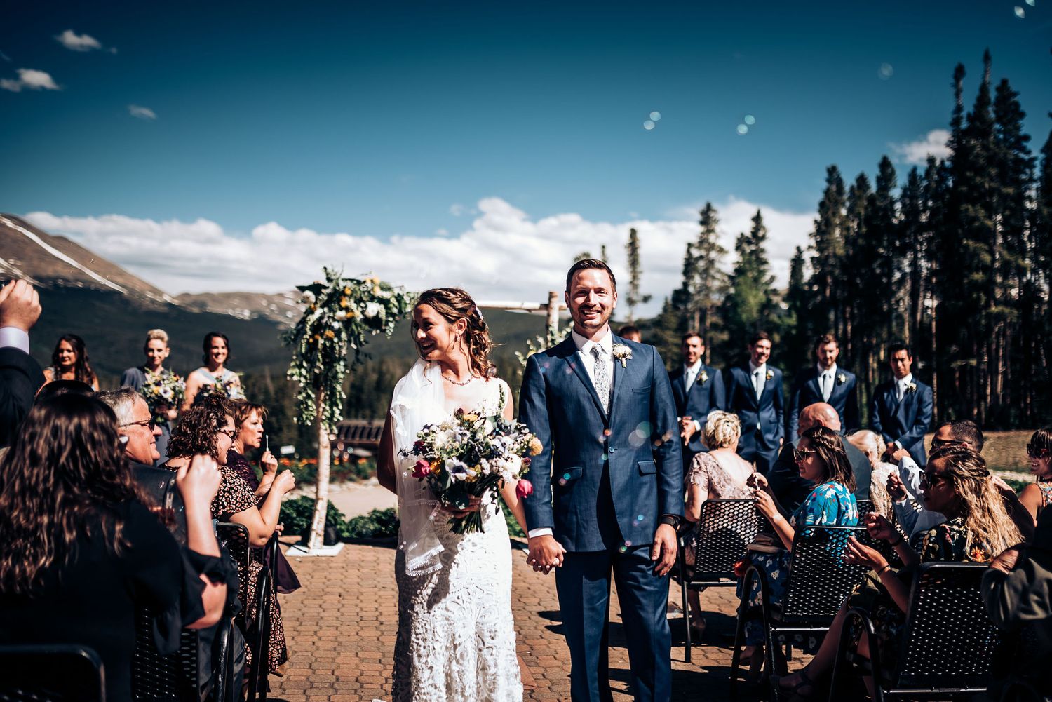 Newlyweds walking down aisle at outdoor mountain wedding while guests celebrate.