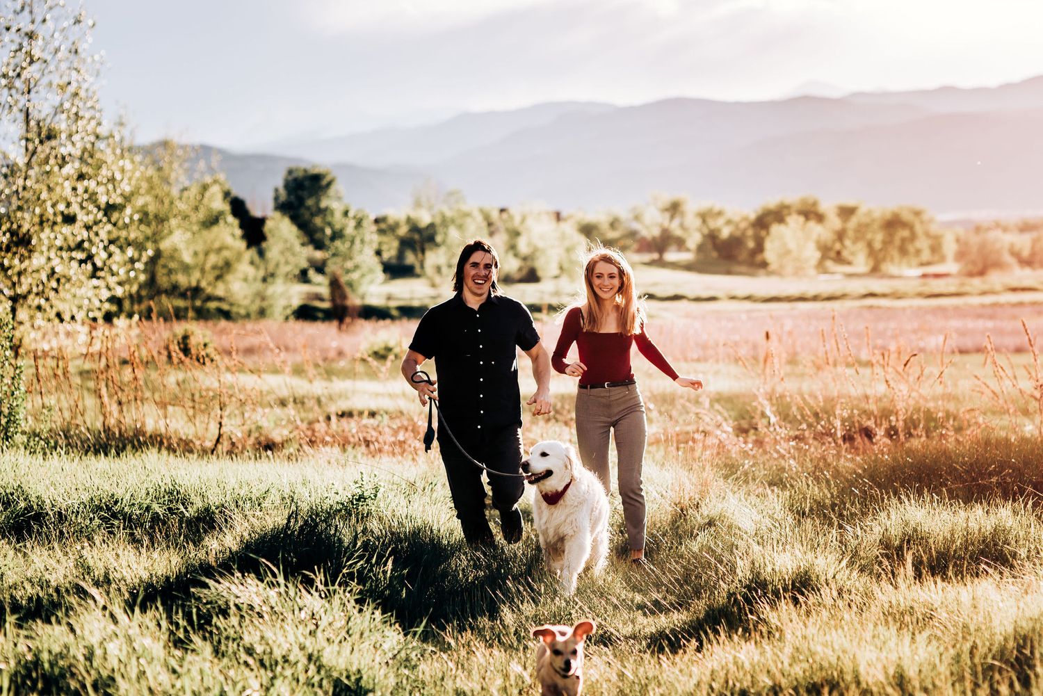 A couple runs through a meadow with their white dog against a mountain backdrop.