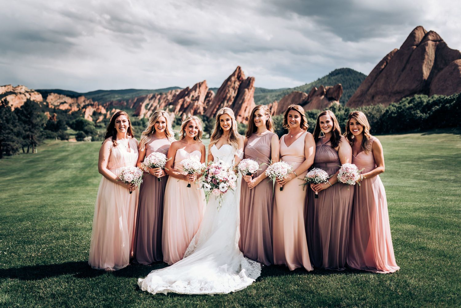 Bridal party in flowing pink dresses posing against red rock formations at garden venue.
