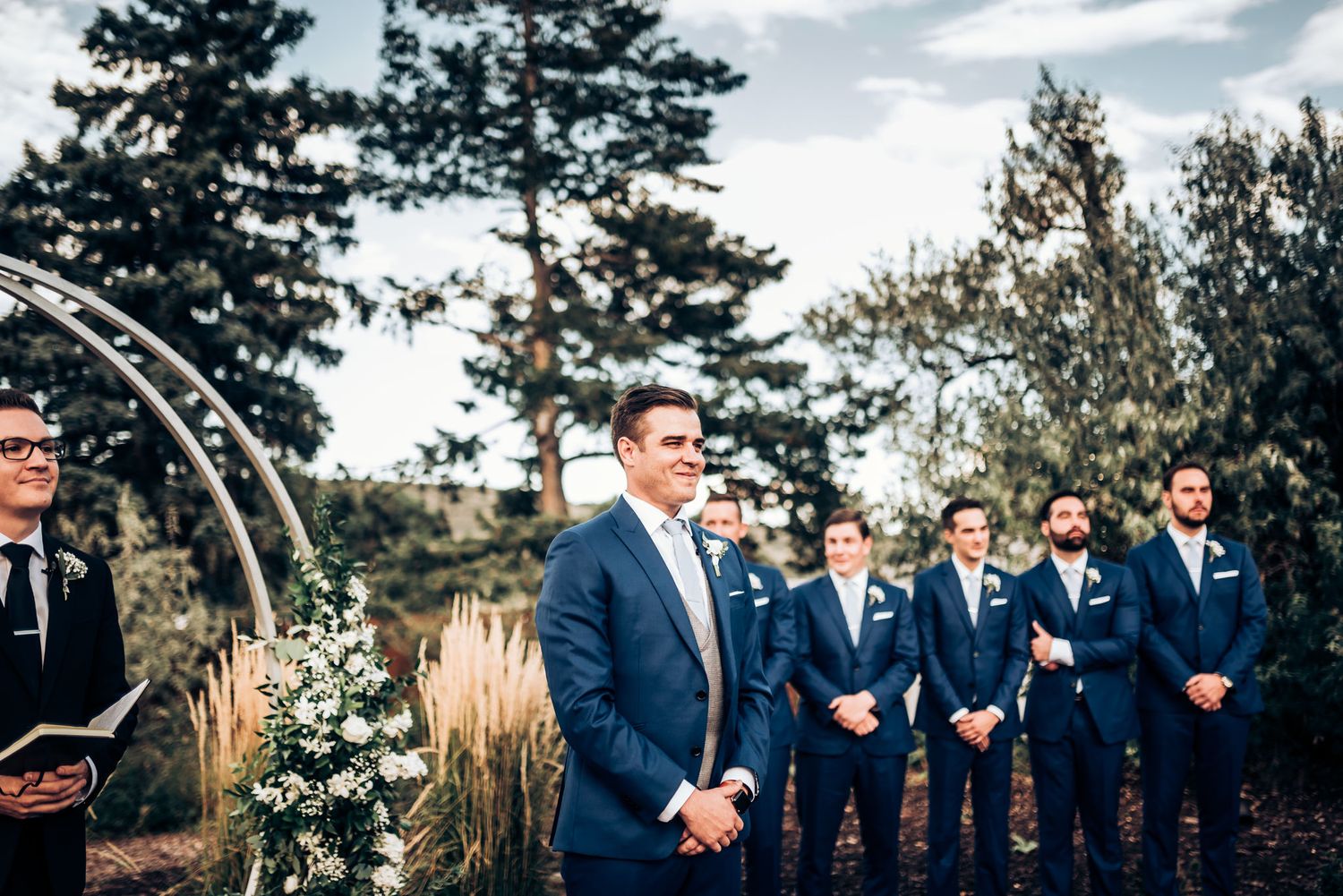 Wedding party lined up at outdoor ceremony site with decorative arch and natural landscaping.