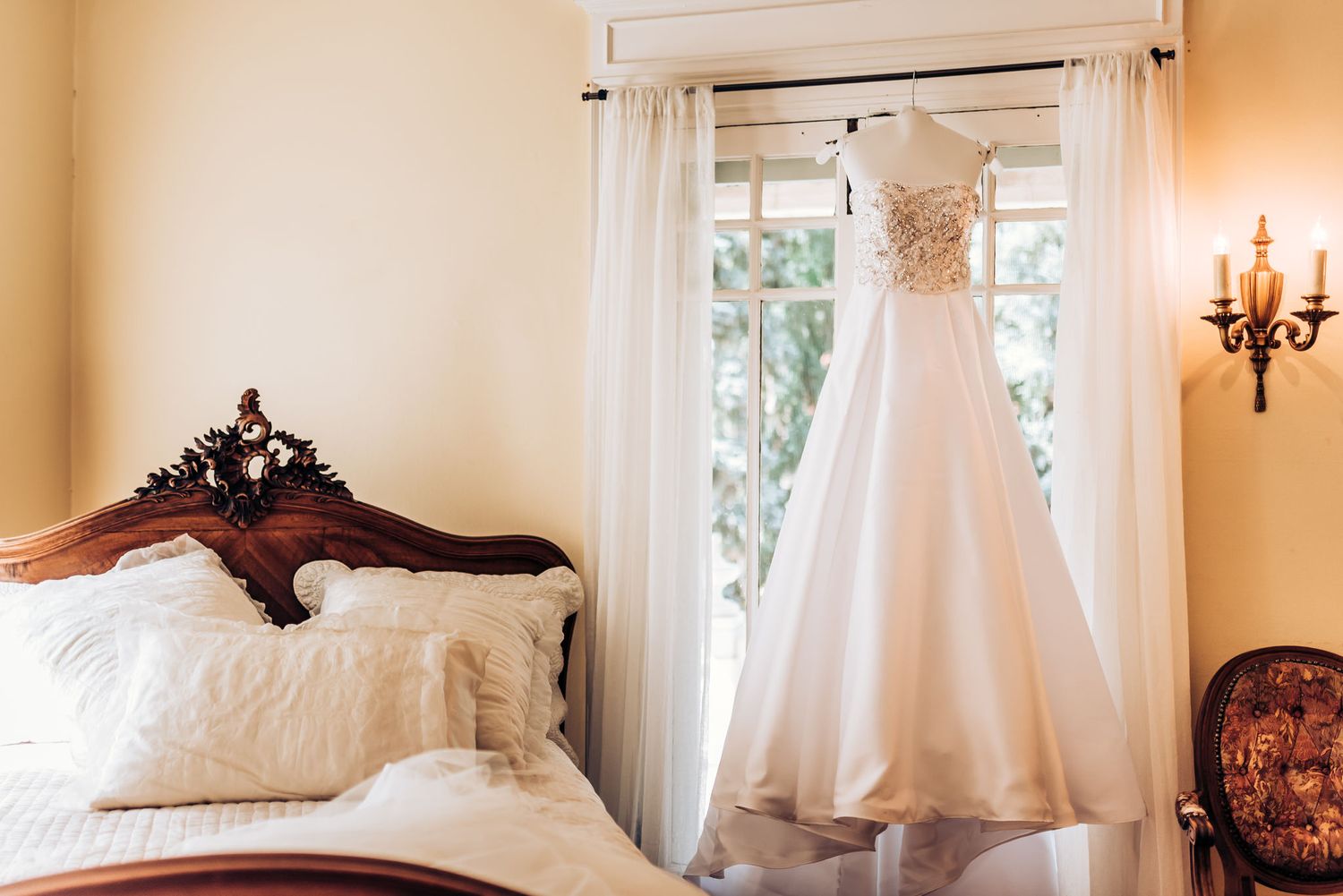 An elegant white wedding gown hangs in a sunlit room with vintage furniture and decor.