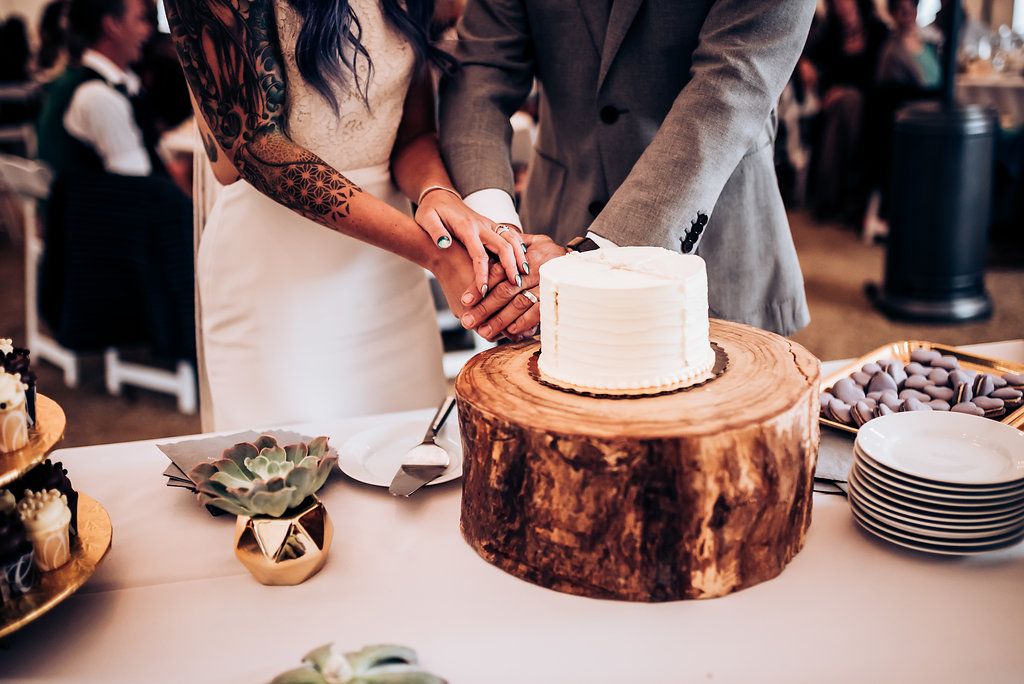 Wedding cake being cut on rustic wood slice cake stand at reception.