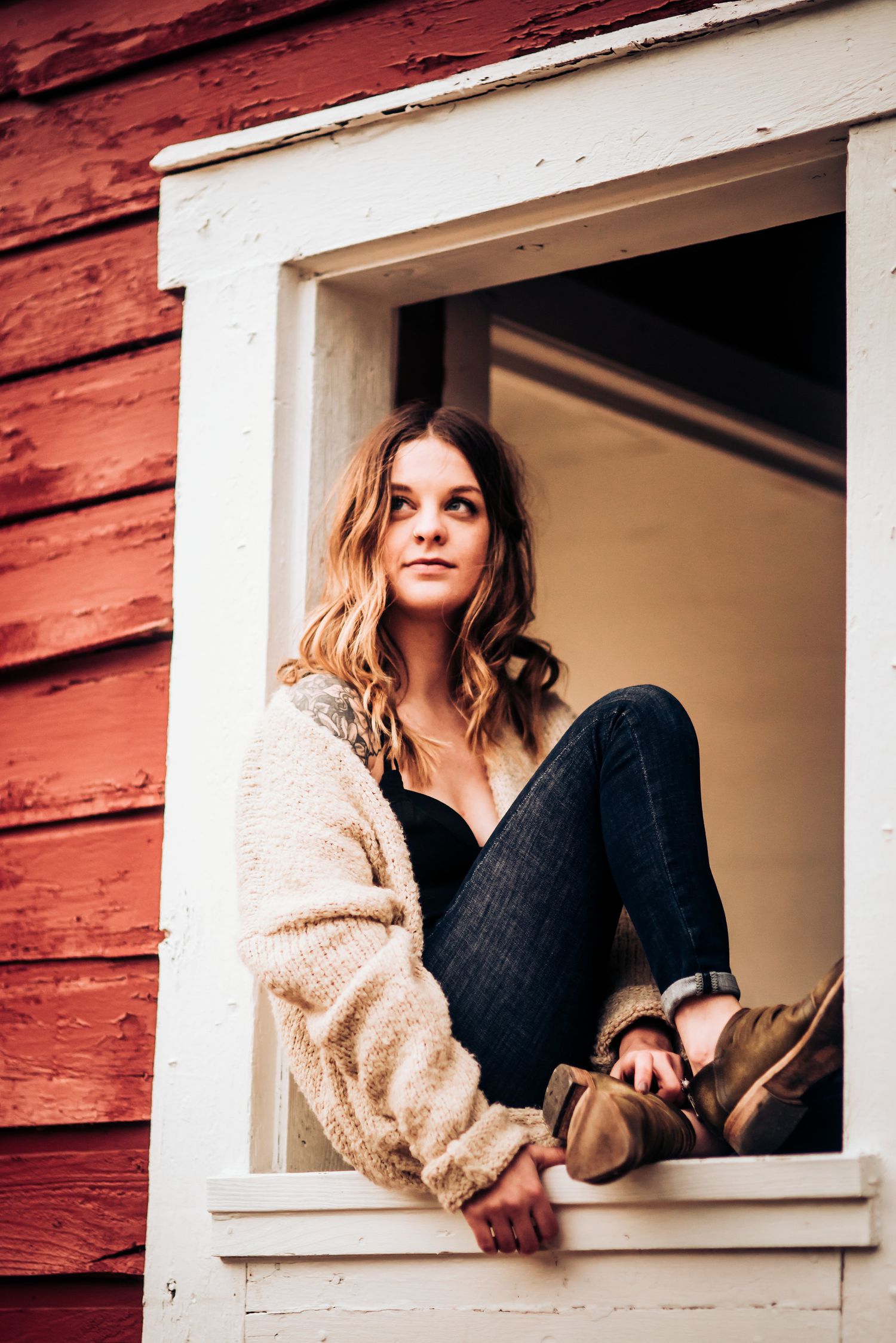 Person sitting in white window frame of red barn wearing cream sweater and jeans.