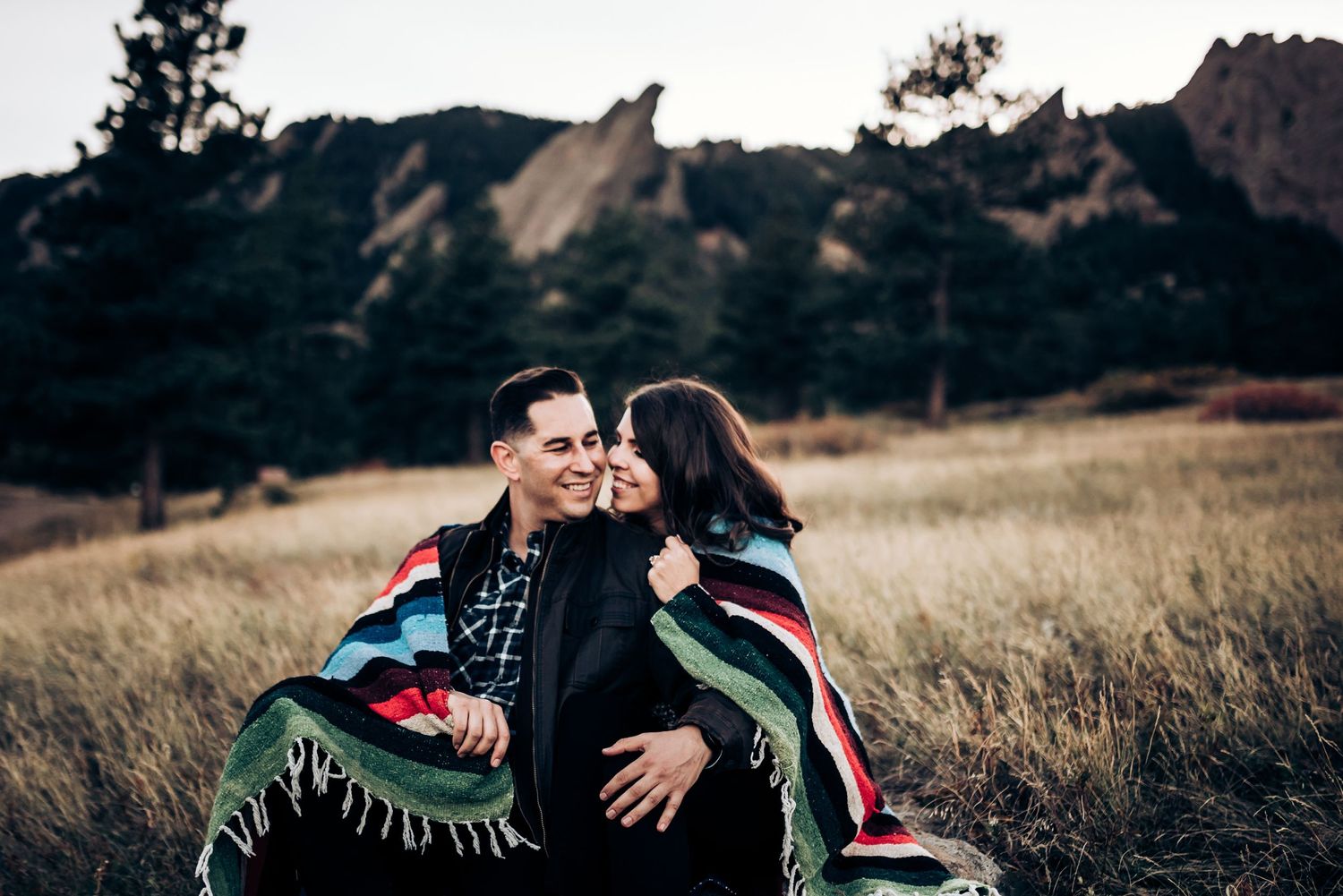 Couple wrapped in a blanket sitting in a field with rocky mountain backdrop.