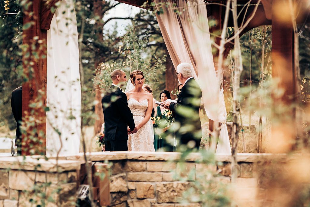 Outdoor wedding ceremony viewed through trees with draped fabric decor.