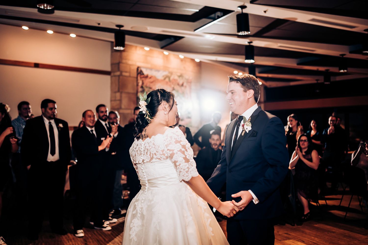 Newlyweds share their first dance in an intimate indoor reception space with exposed beams and decorative lighting.
