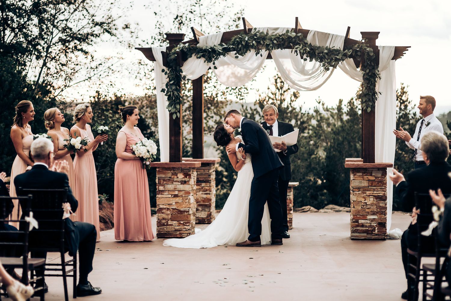 Wedding couple sharing first kiss under rustic arch with stone pillars and draped fabric.