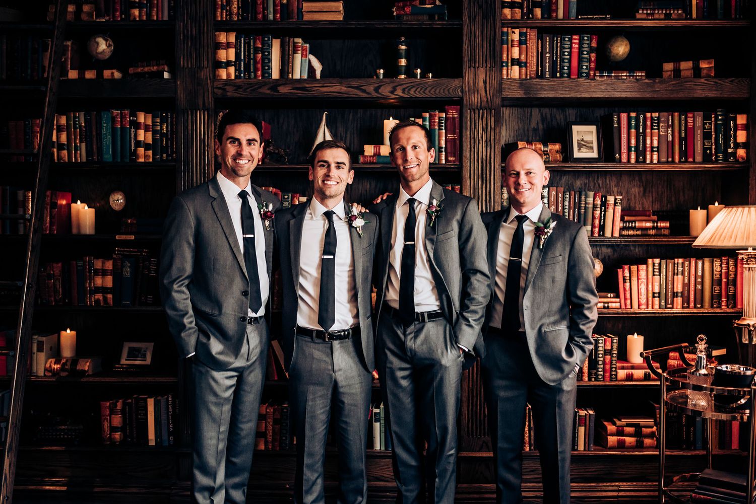 Groomsmen in matching grey suits pose against library bookshelf background.