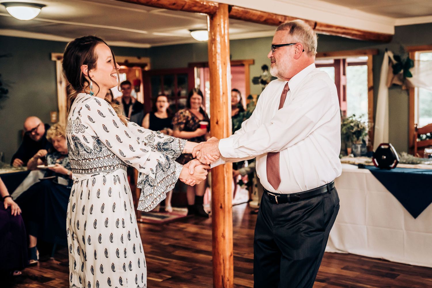 Two people holding hands while dancing at an indoor celebration with wooden support beams.