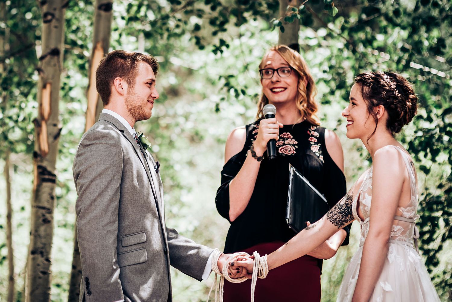 An officiant in burgundy conducts a joyful outdoor wedding ceremony among the trees.