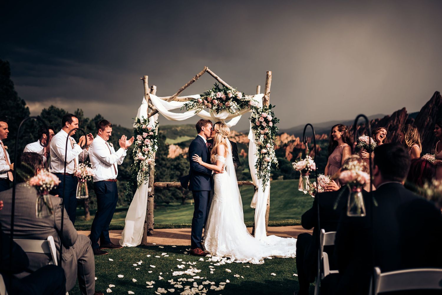 Evening wedding ceremony under white draped arch with scattered rose petals along outdoor aisle.