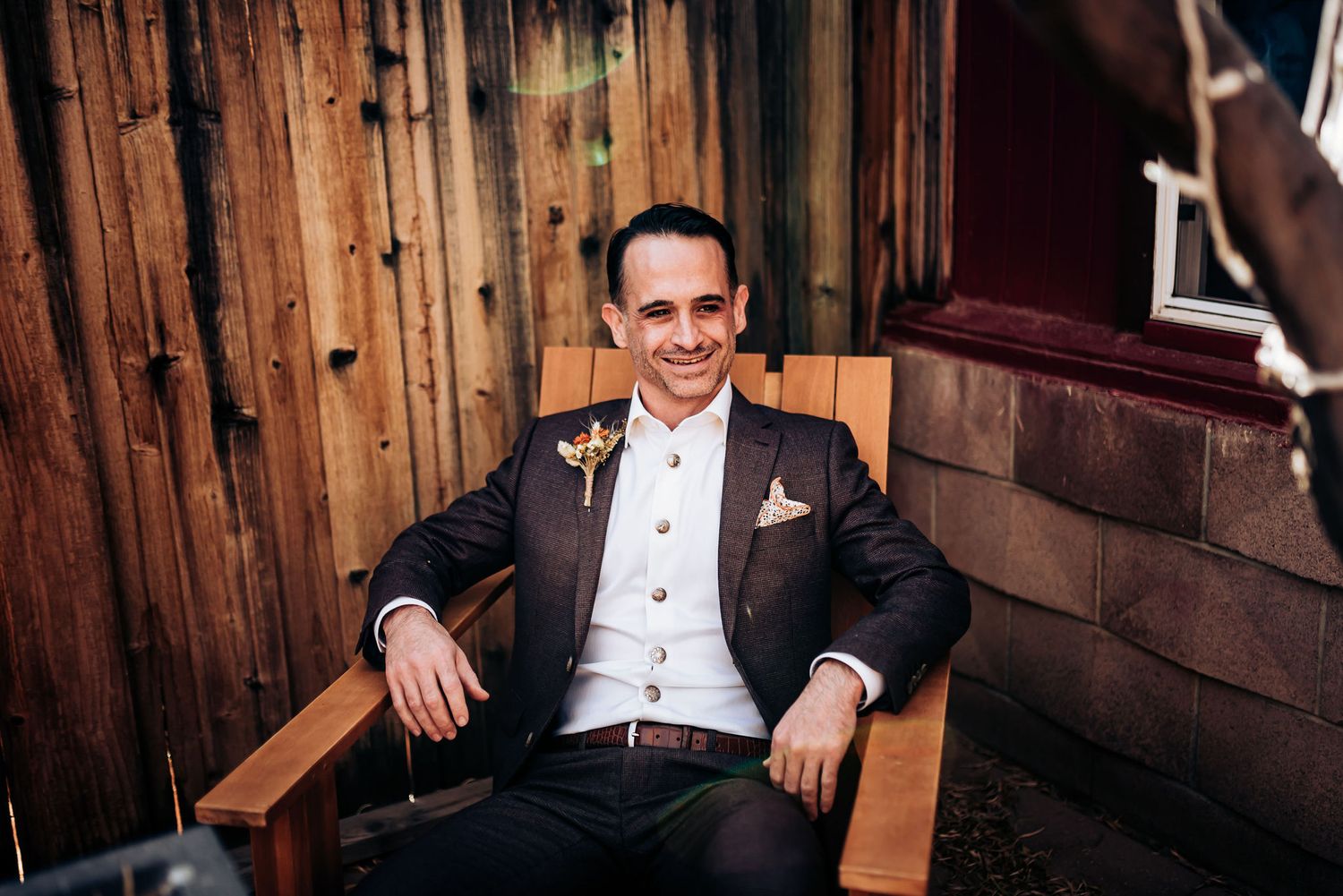 Person in formal attire seated in wooden chair against rustic wall.