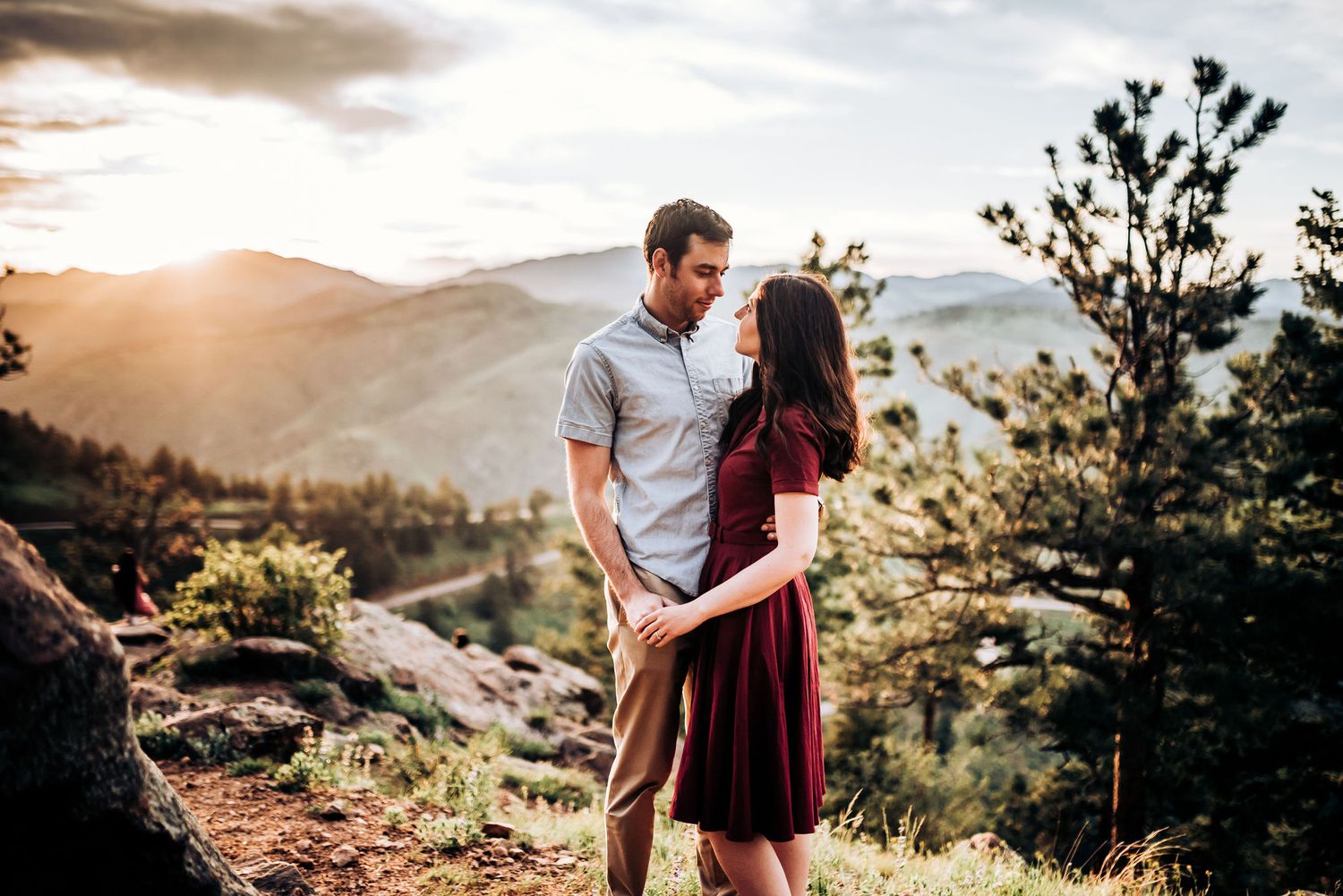 Couple shares tender moment on mountain overlook with sunset-lit valley behind them.