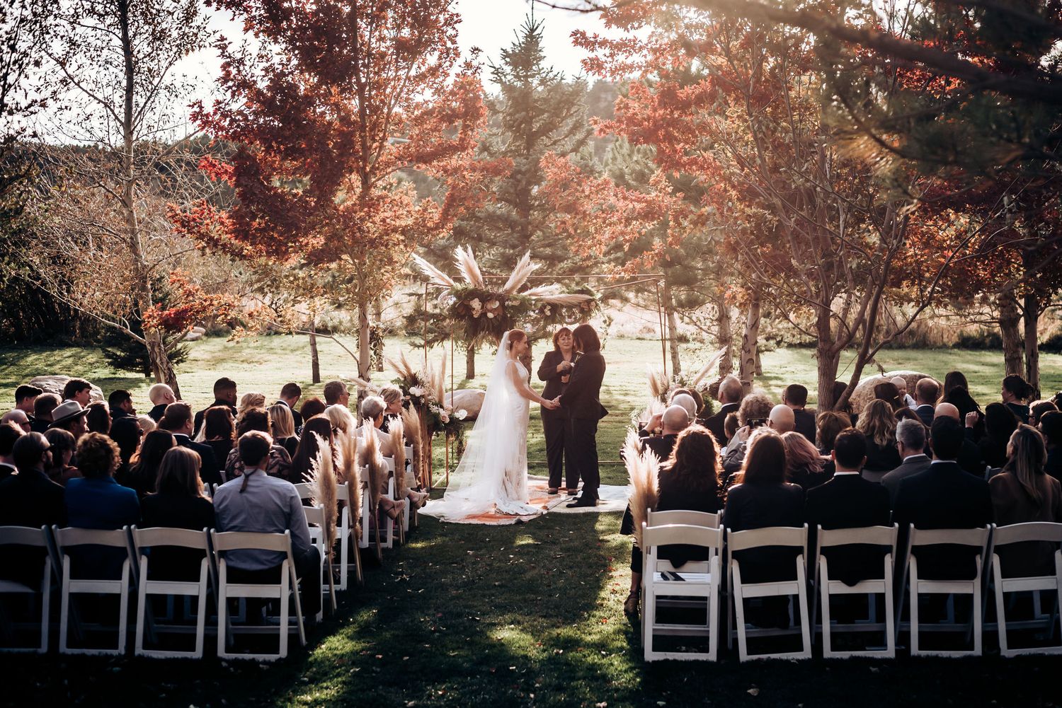 Wide view of an outdoor fall wedding ceremony with white chairs and many guests in attendance.