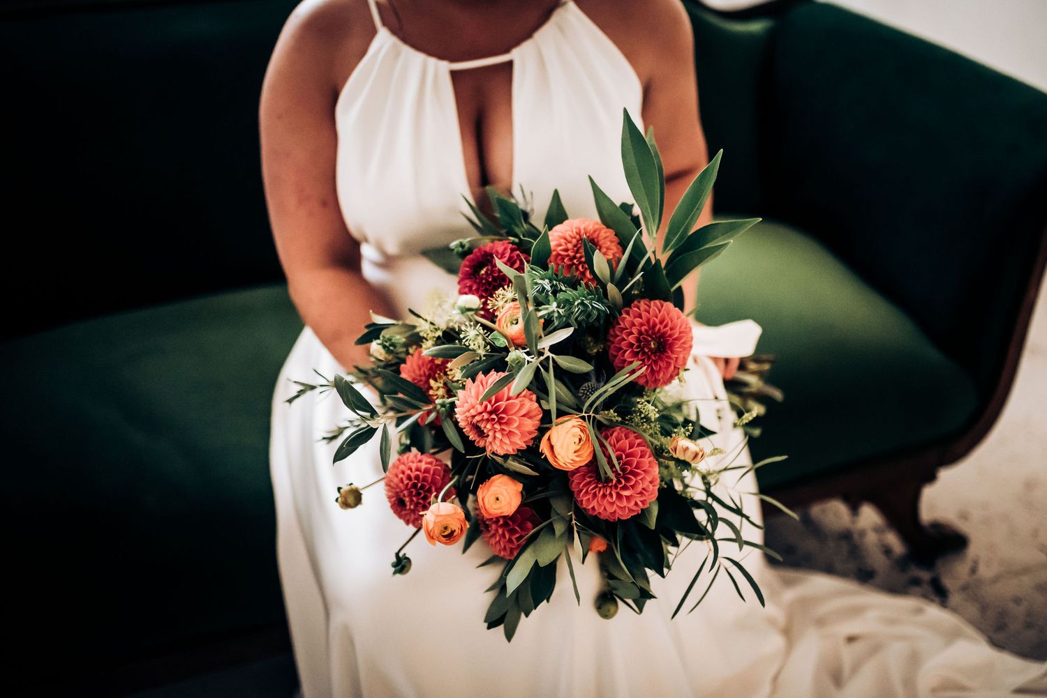 Vibrant wedding bouquet with coral dahlias and greenery displayed against emerald velvet upholstery.