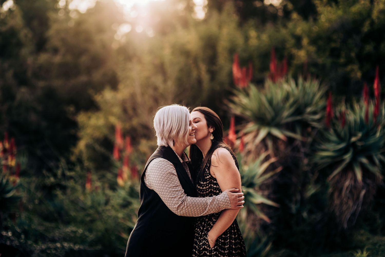 A loving moment captured in a garden with red flowering plants and warm backlighting.
