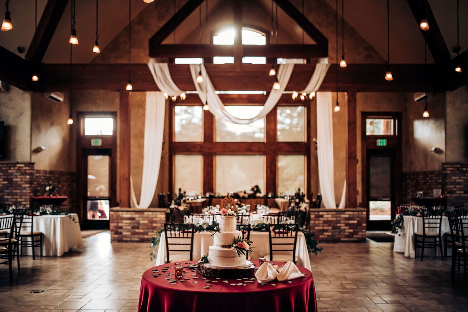 Wedding reception venue with vaulted wooden ceiling, white drapes, and burgundy accents.