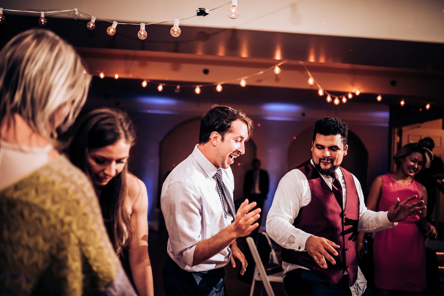 Party guests dancing and enjoying themselves at a wedding reception with string lights overhead.