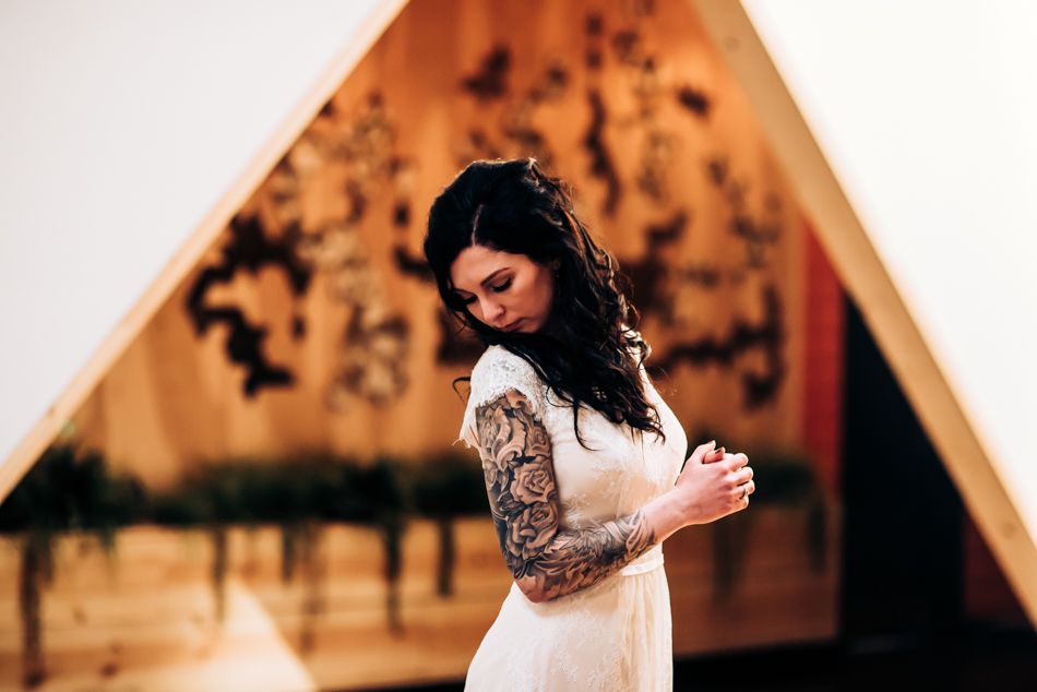 A bride in a white dress with intricate sleeve tattoos stands in soft lighting against a warm backdrop.