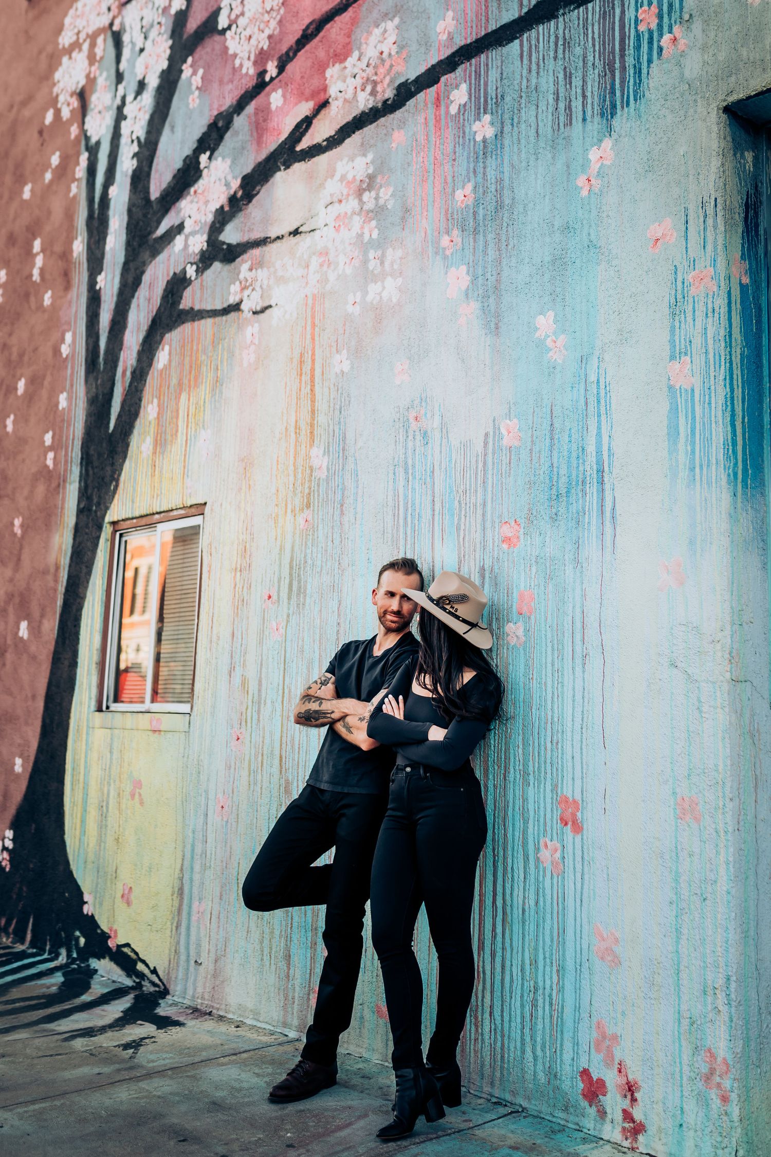 A couple stands against a turquoise wall with painted tree branches and pink blossoms.