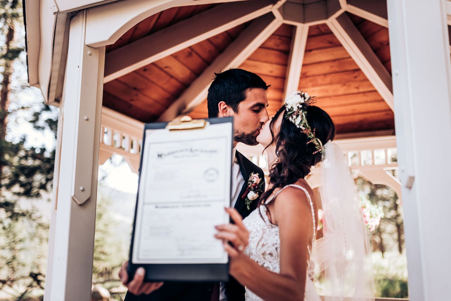 A couple kisses while displaying their marriage certificate in a wooden gazebo.