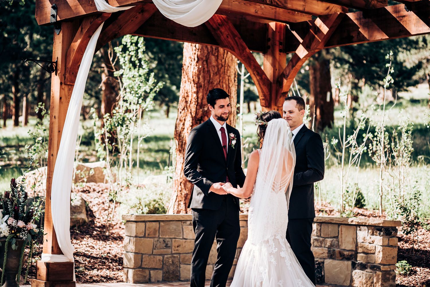 A wedding ceremony unfolds beneath a wooden pergola with stone pillars in a forest setting.