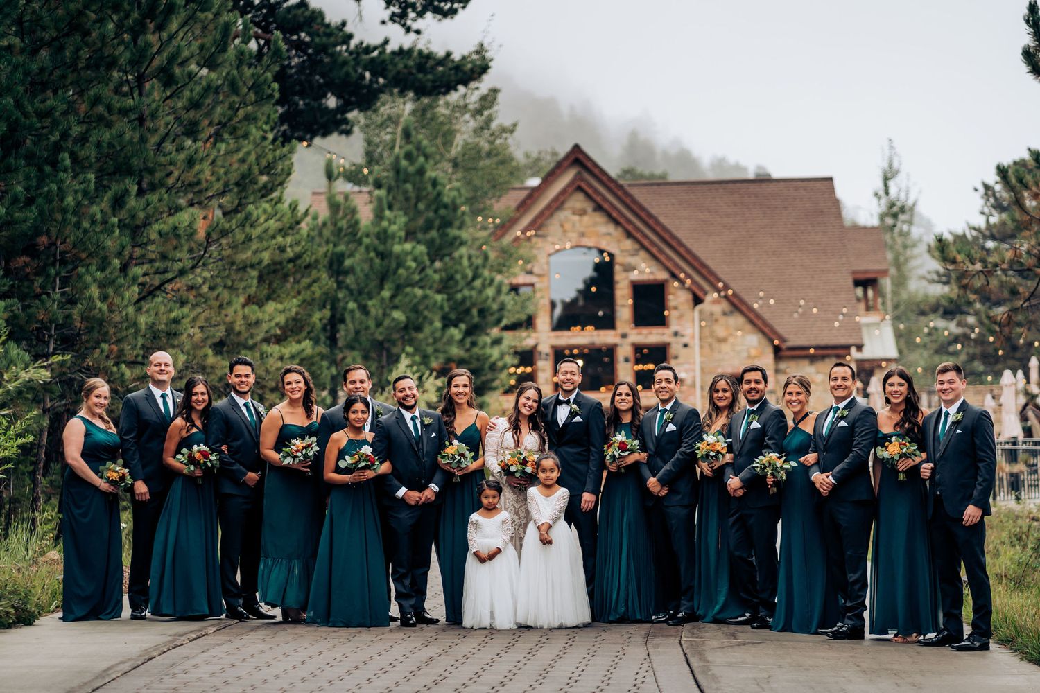 Large wedding party in teal dresses and navy suits poses in front of rustic wooden lodge.