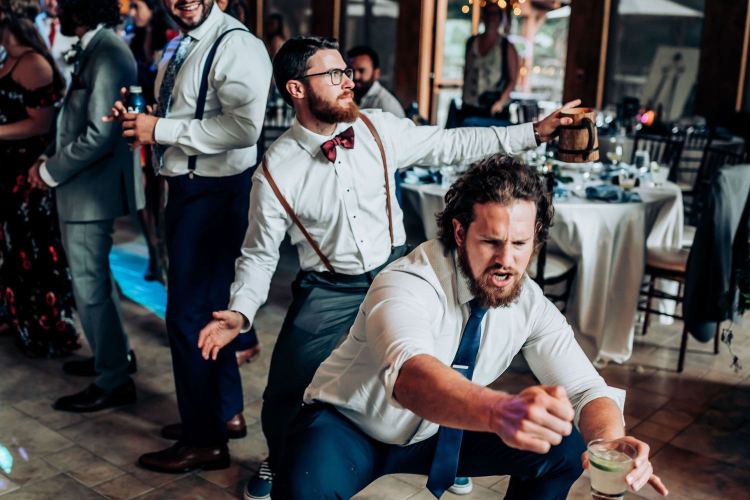 Wedding guests in suspenders and bow ties energetically dance during the reception celebration.