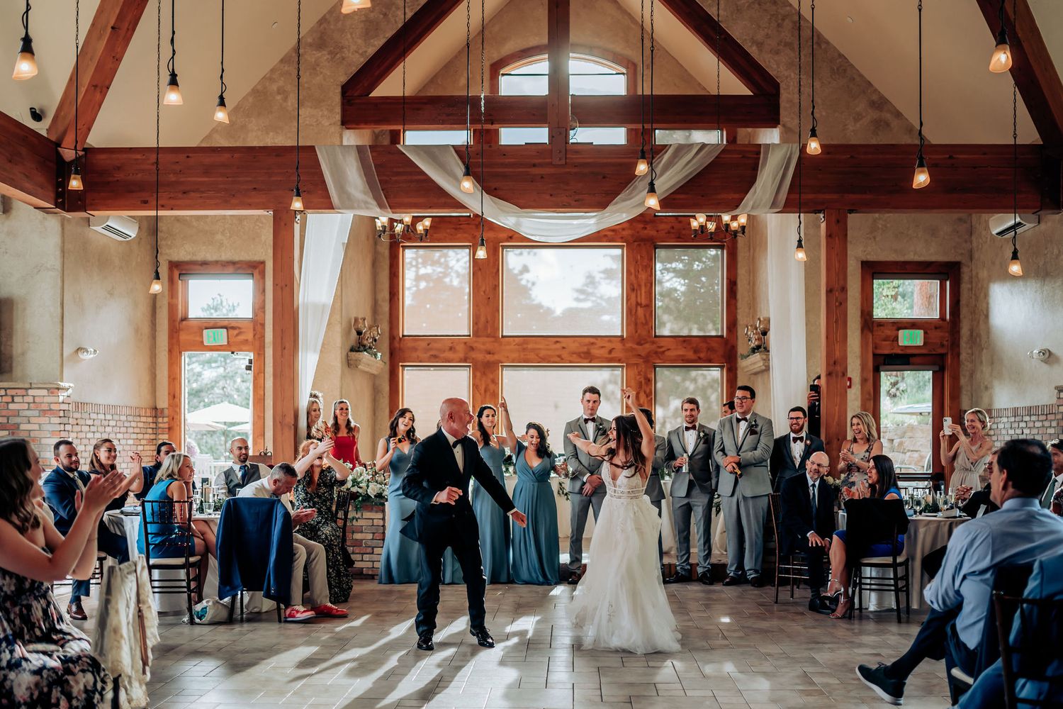 Wedding party dances in a spacious rustic venue with wooden beams and natural lighting.