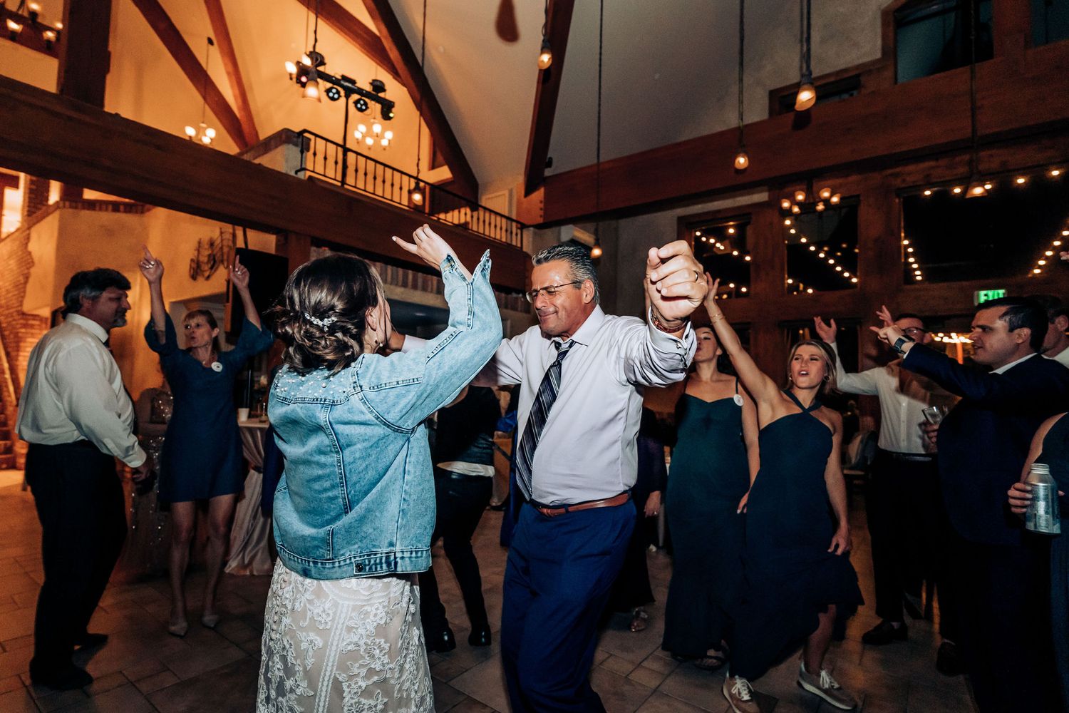 Party guests dancing energetically at a wedding reception in a rustic venue with wooden beams.