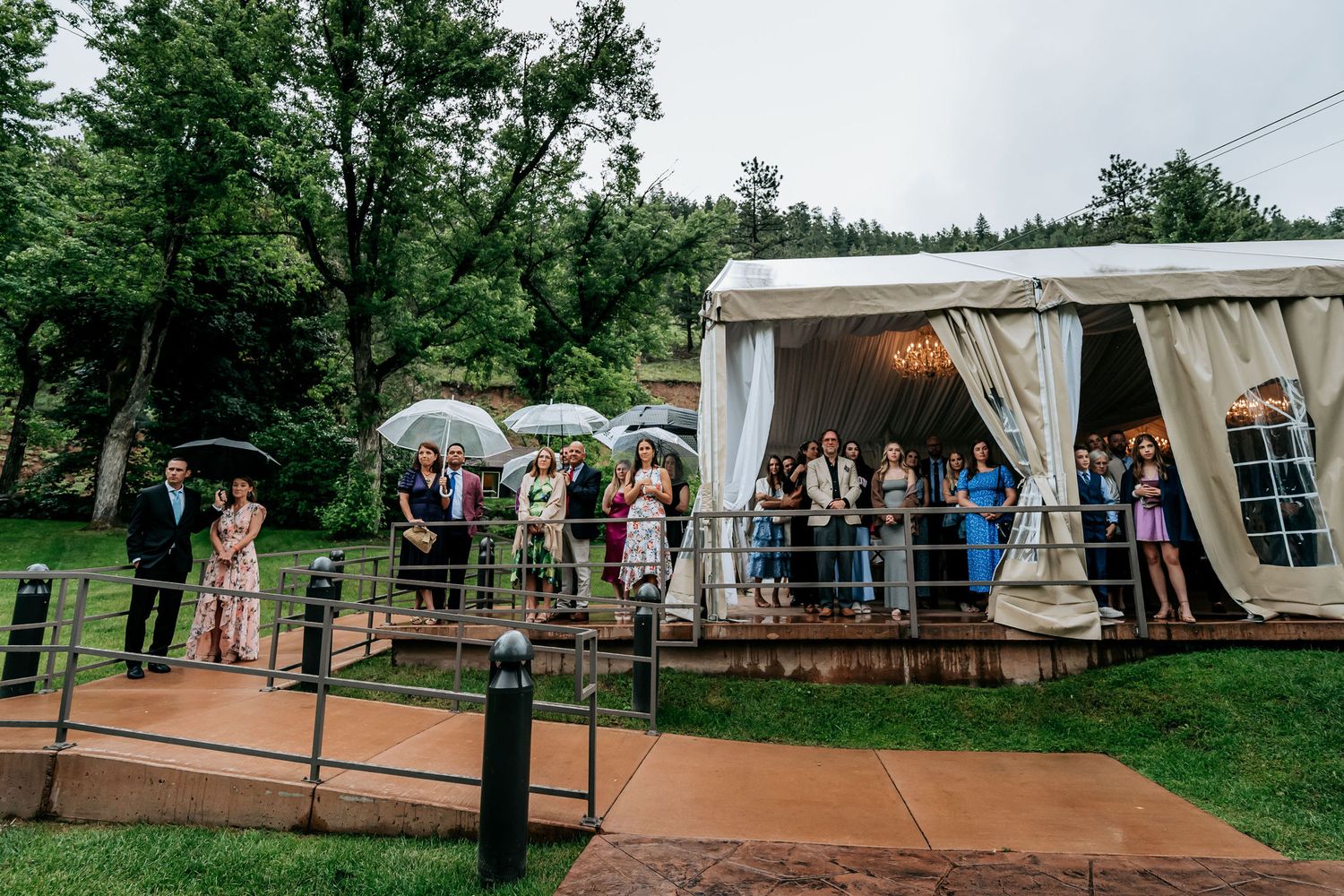 Wedding reception tent setup with guests gathering on wooden deck surrounded by green forest.