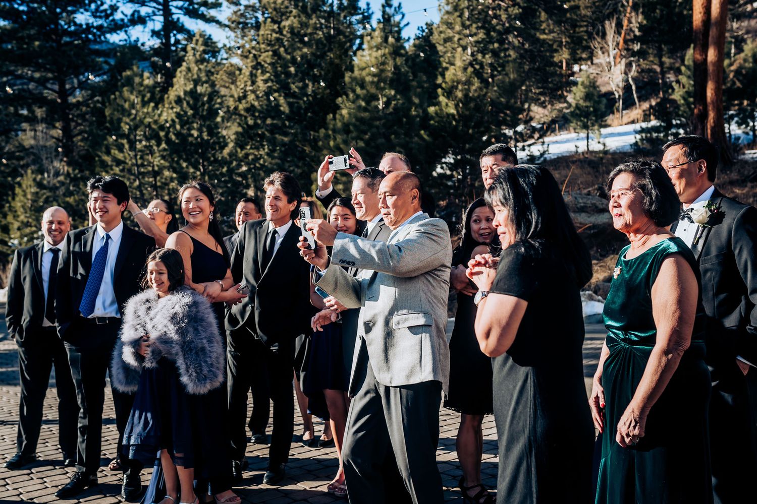 Large wedding party gathered outdoors on paved terrace with mountain scenery in background.