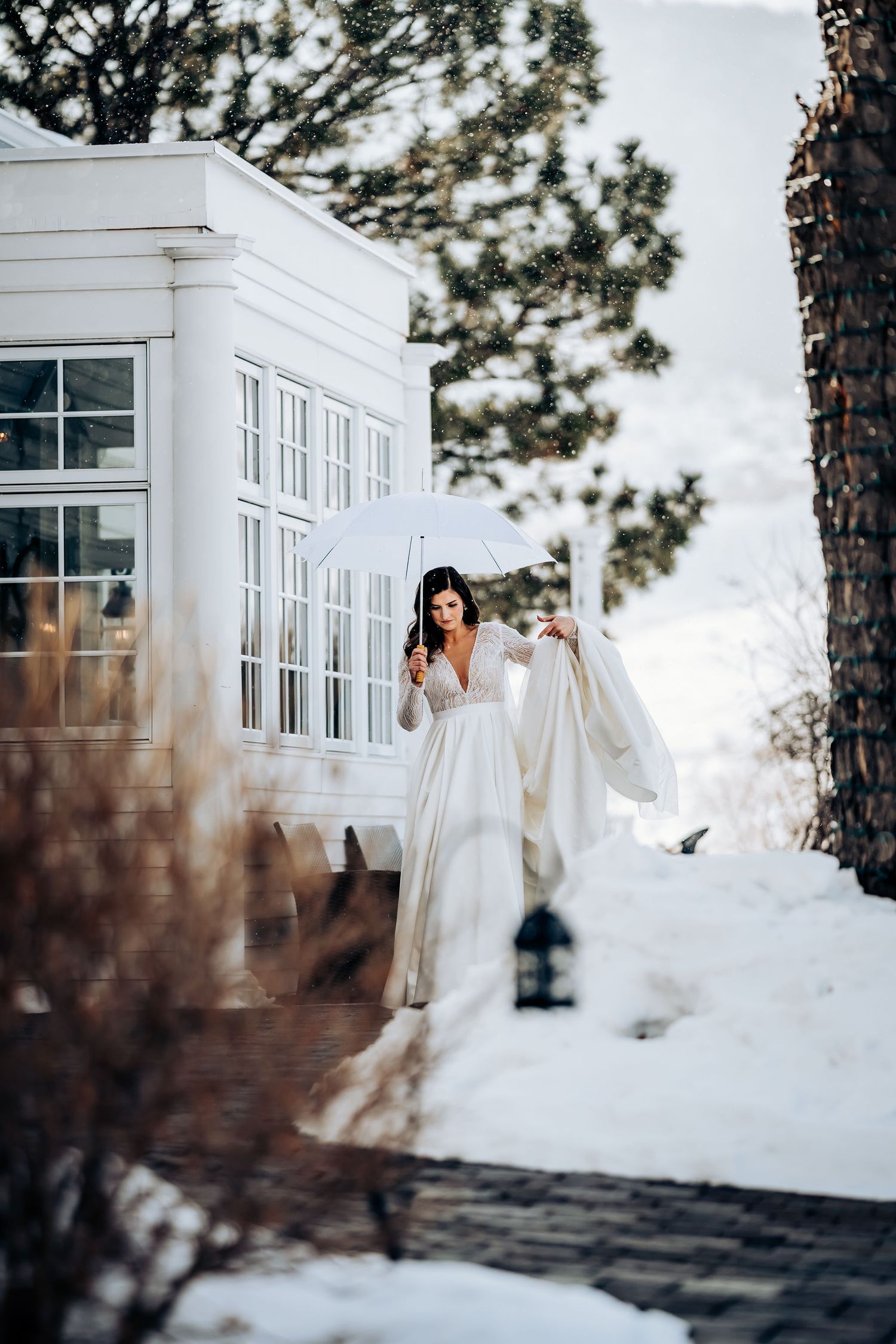 A white wedding dress flows in the winter breeze against a snowy backdrop and white colonial building.