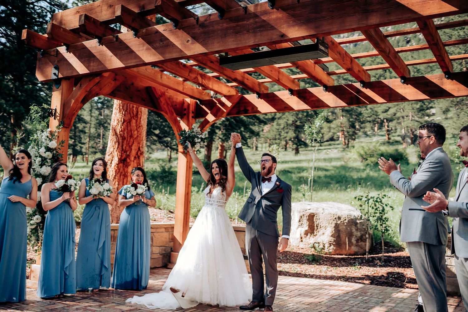 Wedding celebration with wedding party in blue dresses beneath wooden pergola during outdoor ceremony.