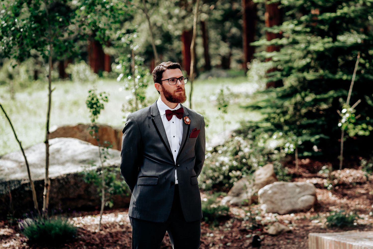 A groom in a dark suit and bow tie poses in an outdoor woodland setting.