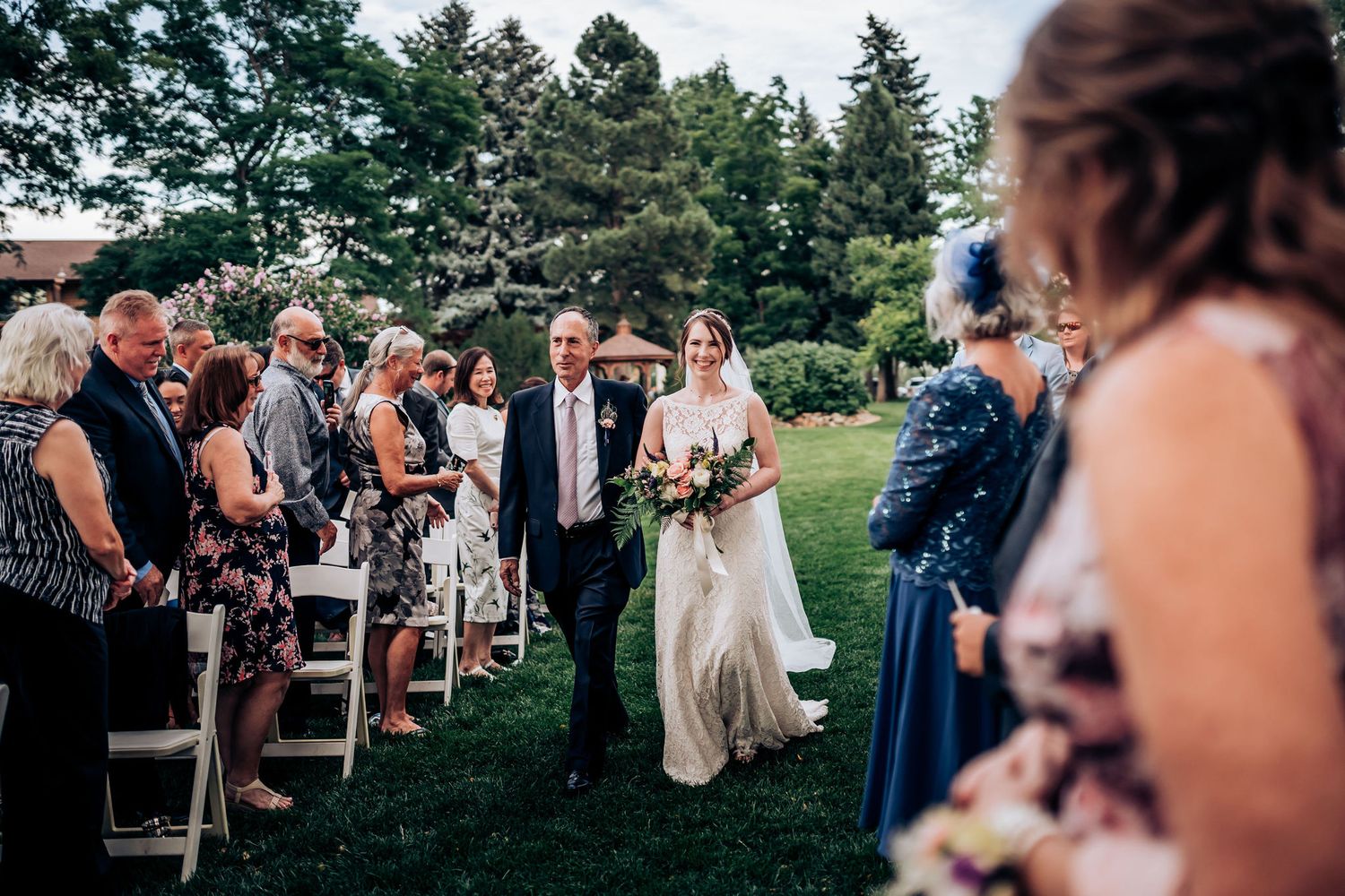 Wedding ceremony aisle with guests seated on white chairs outdoors among evergreen trees.