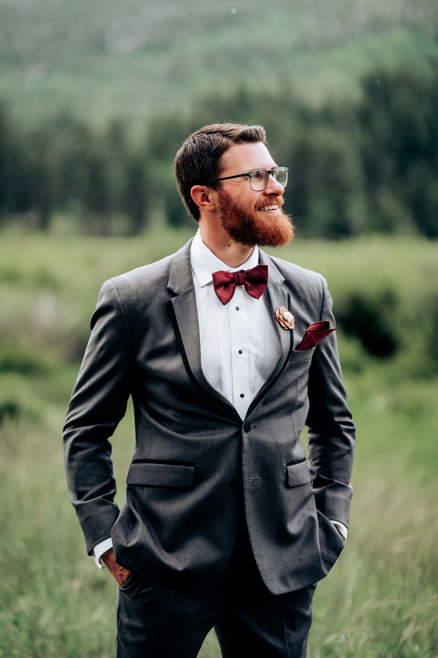 Groom in grey suit and burgundy bow tie smiles in an outdoor mountain setting.