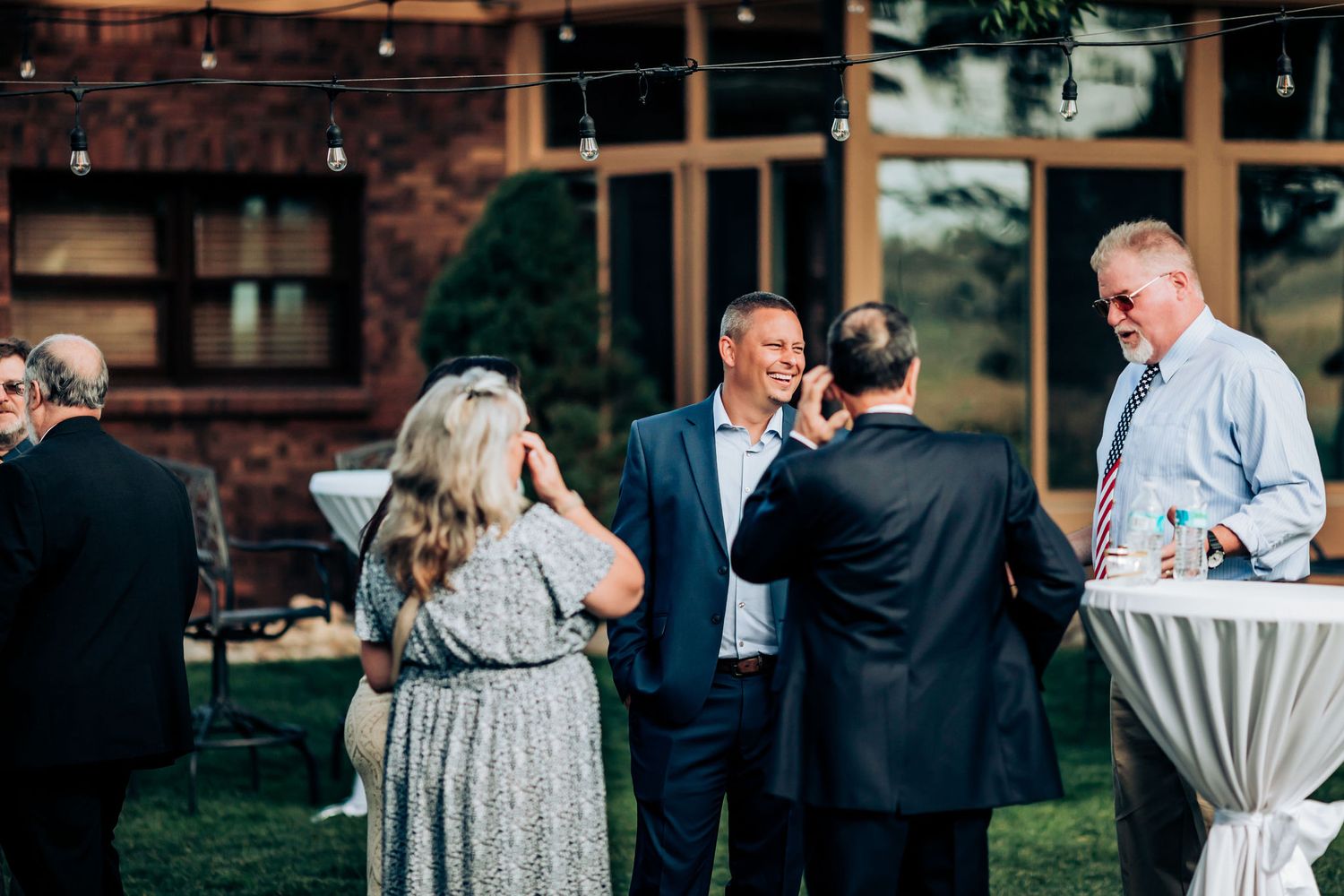 Wedding guests mingle and socialize at an outdoor cocktail hour with high-top tables on a manicured lawn.
